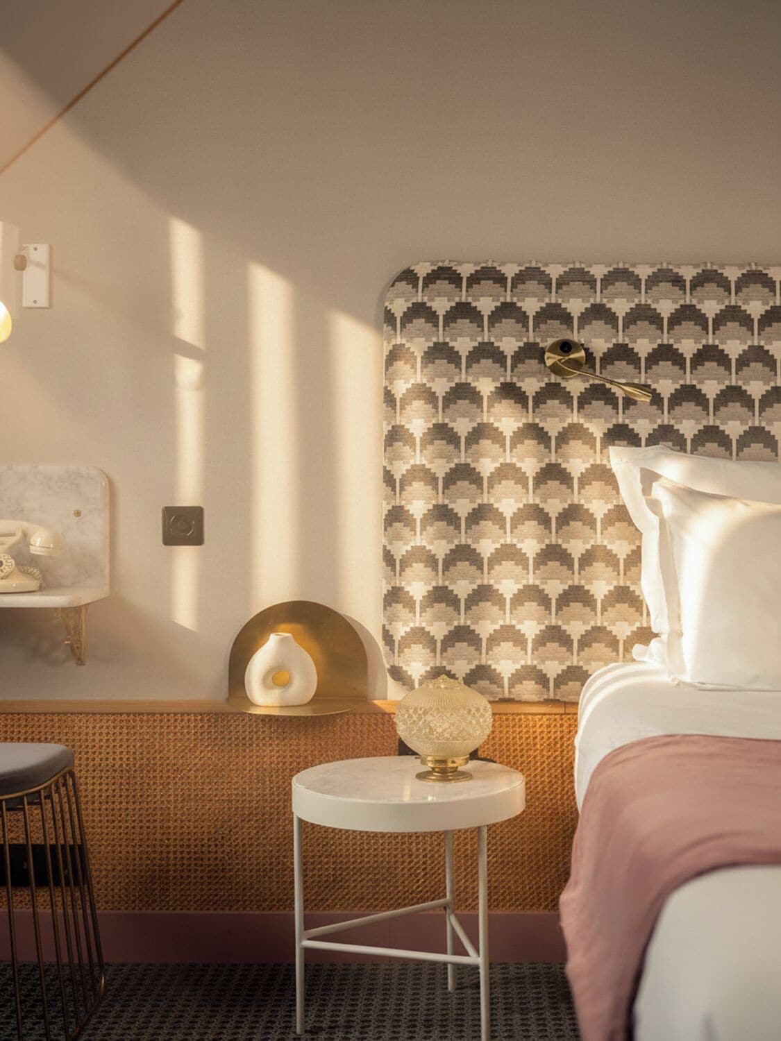 A modern hotel room with a patterned wallpaper, a round table with a glass top, a classic telephone, and a bed with white linens.