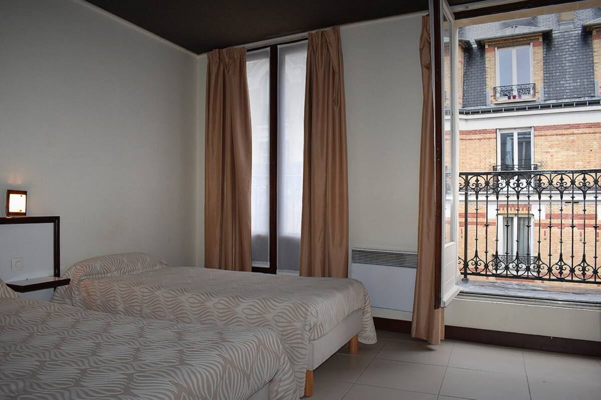 A bedroom with a bed, a window with curtains, a radiator, and a view of a balcony with a fence.