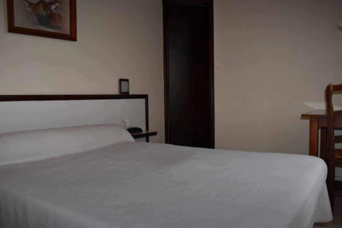 A simple, unadorned hotel room with a single bed, a wooden chair, and a small bedside table.
