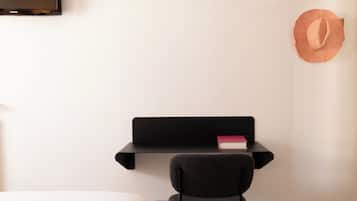 Una habitación con una cama, una silla negra, un estante negro con un libro y un sombrero colgado en la pared.
