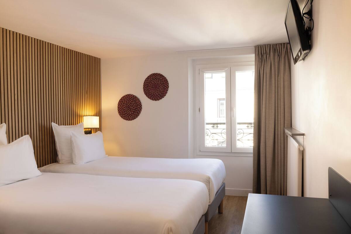 A hotel room with two beds, a TV mounted on the wall, a window with curtains, and a balcony view.