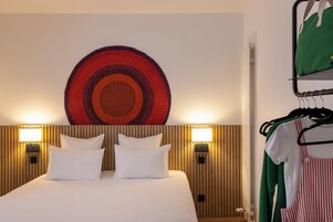 A hotel room with a bed, two wall-mounted lamps, a wall art piece, and a clothing rack with hangers.