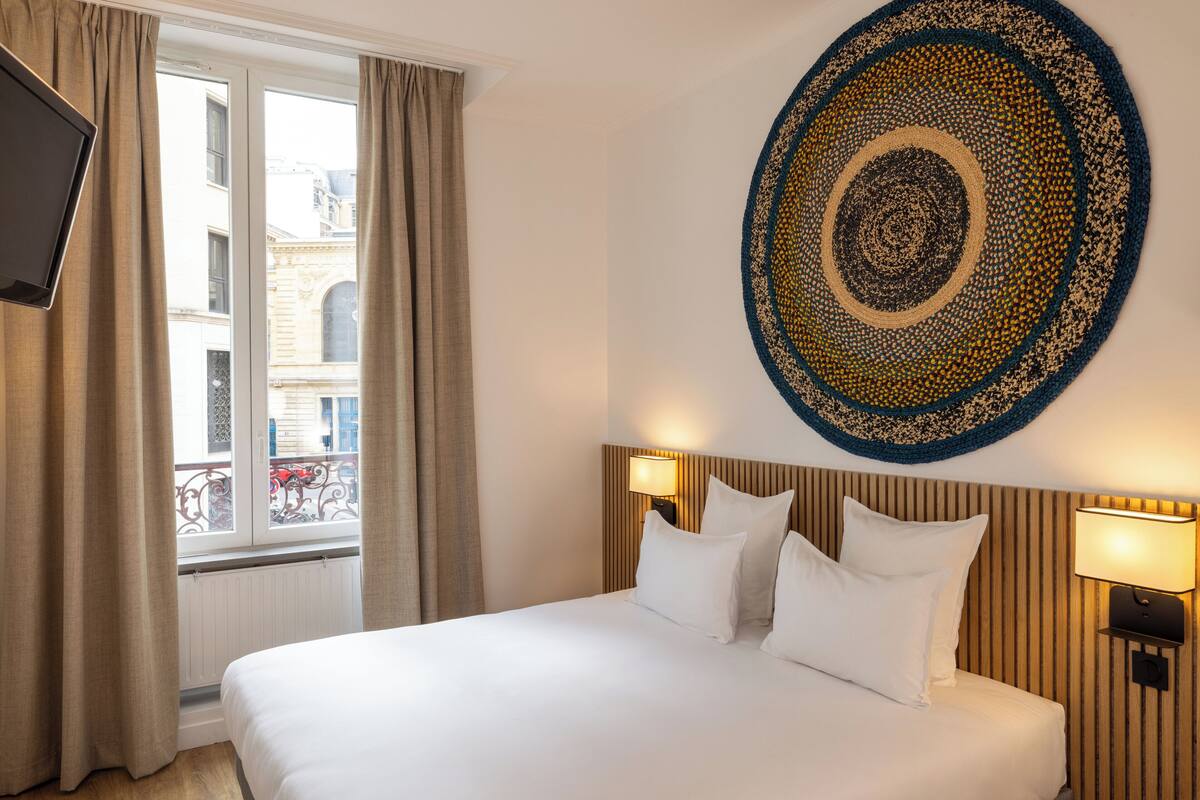A hotel room with a bed, two bedside lamps, a wall-mounted TV, and a large decorative rug.
