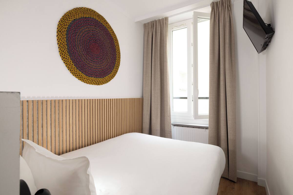 A hotel room with a bed, a wall art piece, a window with curtains, and a mounted TV.