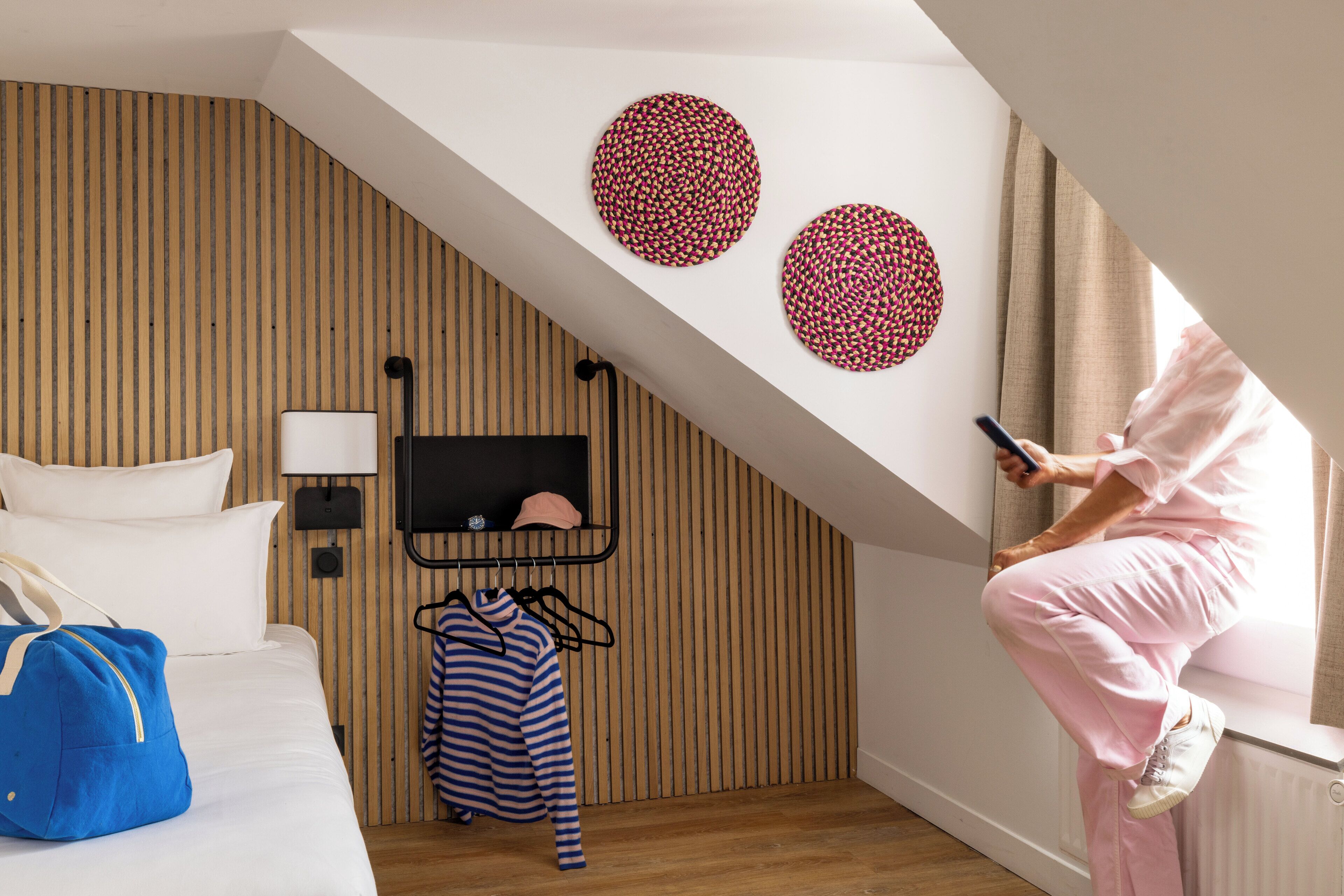 Habitación de hotel con techo inclinado, una cama con ropa blanca, un bolso azul sobre la cama, un perchero con prendas a rayas y una persona usando un teléfono inteligente.