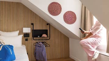 Habitación de hotel con techo inclinado, una cama con ropa blanca, un bolso azul sobre la cama, un perchero con prendas a rayas y una persona usando un teléfono inteligente.