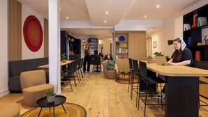 A modern lounge area with seating arrangements, a central table, and a visible kitchen area in the background. - Hôtel Korner Opéra (Paris)