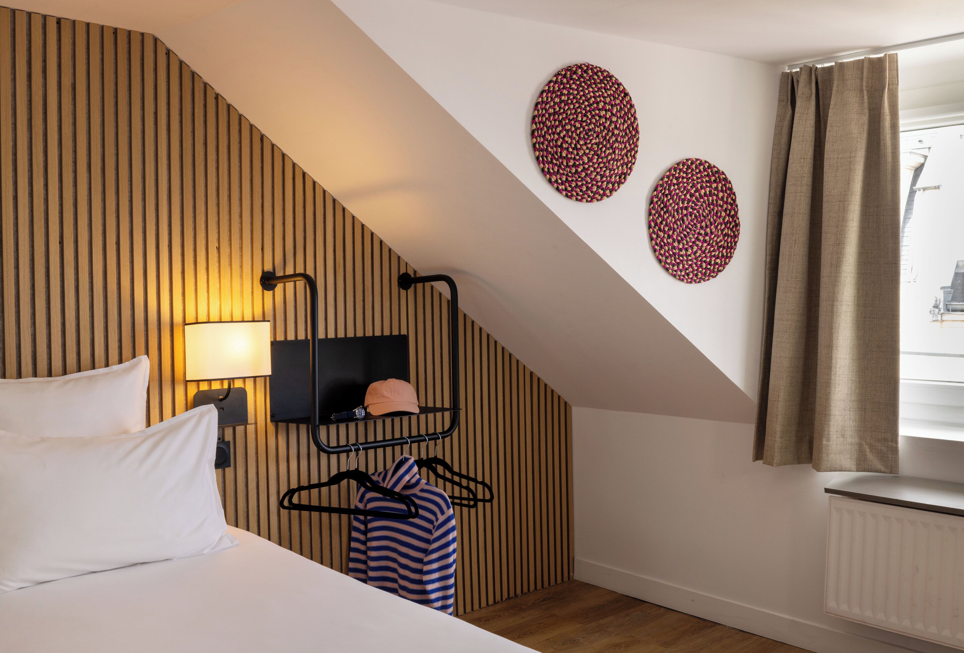 Ein Hotelzimmer mit schrägen Wänden, einem Bett mit weißer Bettwäsche, einem an der Wand befestigten Fernseher, einem Heizkörper und einem mit Vorhängen versehenen Fenster.