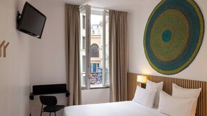 A hotel room with a bed, a TV mounted on the wall, a desk with a chair, and a window with curtains. - Hôtel Korner Opéra (Paris)