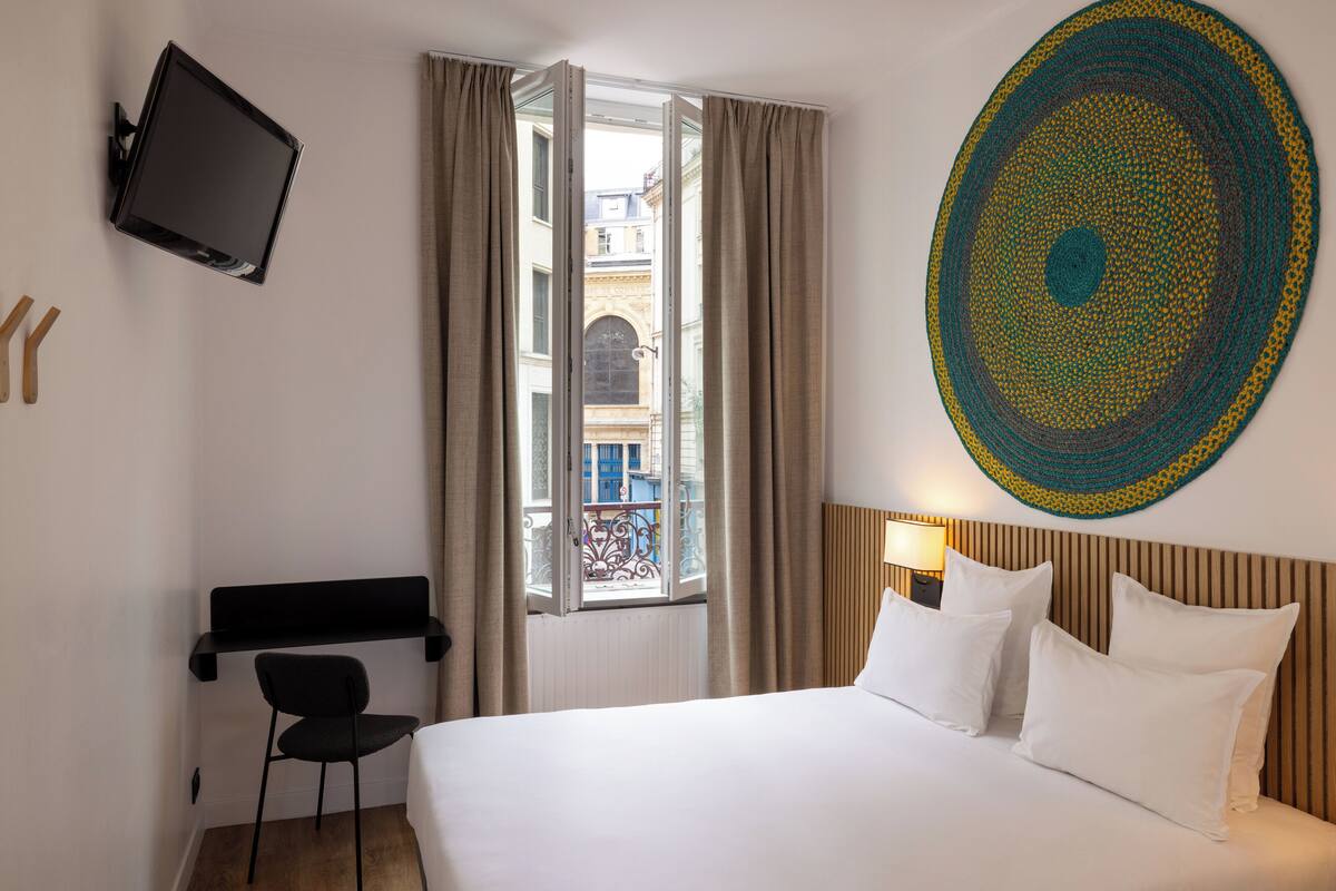 A hotel room with a bed, a TV mounted on the wall, a desk with a chair, and a window with curtains.
