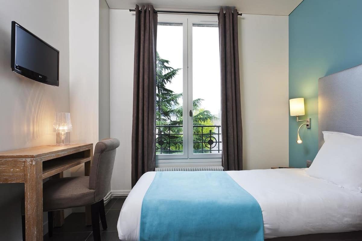 A hotel room with a bed, a desk with a chair, a TV, a window with curtains, and a balcony view.