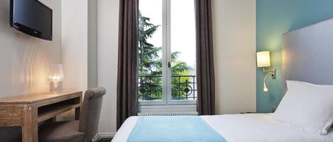 A hotel room with a bed, a desk with a chair, a TV, a window with curtains, and a balcony view.