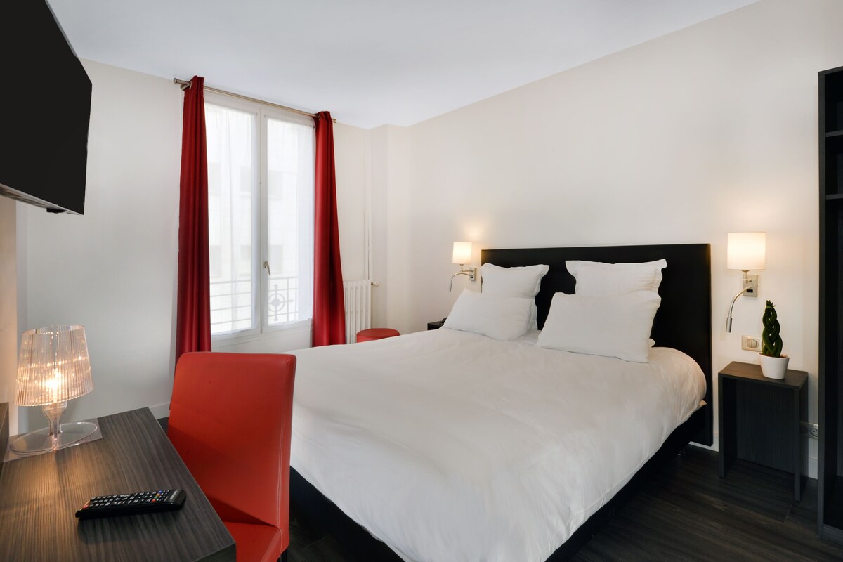 A hotel room with a large bed, red chair, bedside tables with lamps, and a TV on a stand.