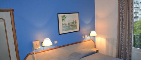 Een hotelkamer met een blauwe muur, een bed met twee kussens, een nachtkastje met een lamp, een lijst met een foto aan de muur en een raam met gordijnen.