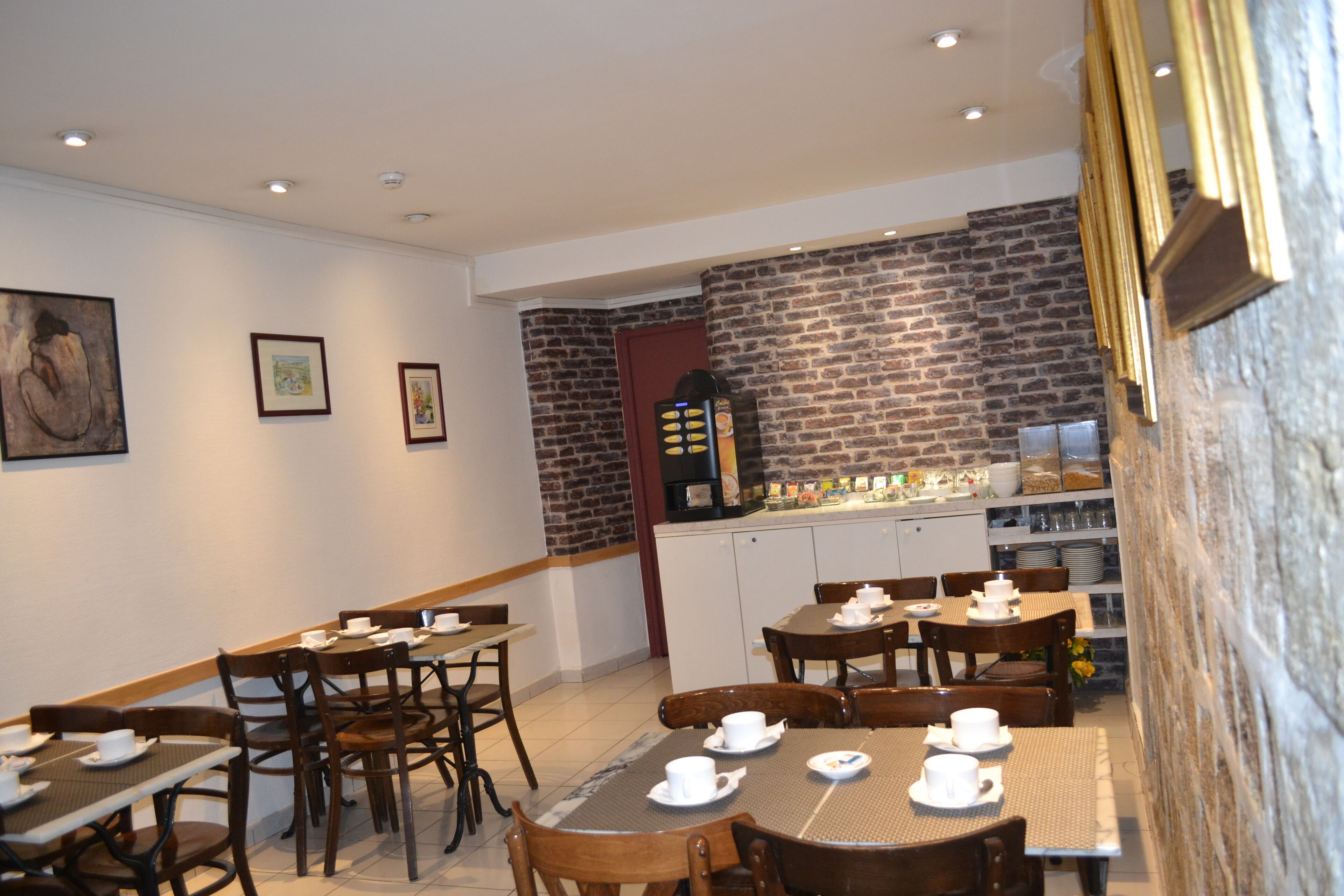 A dining area with brick walls, wooden tables, and chairs. There are framed pictures on the wall and a coffee machine in the background.