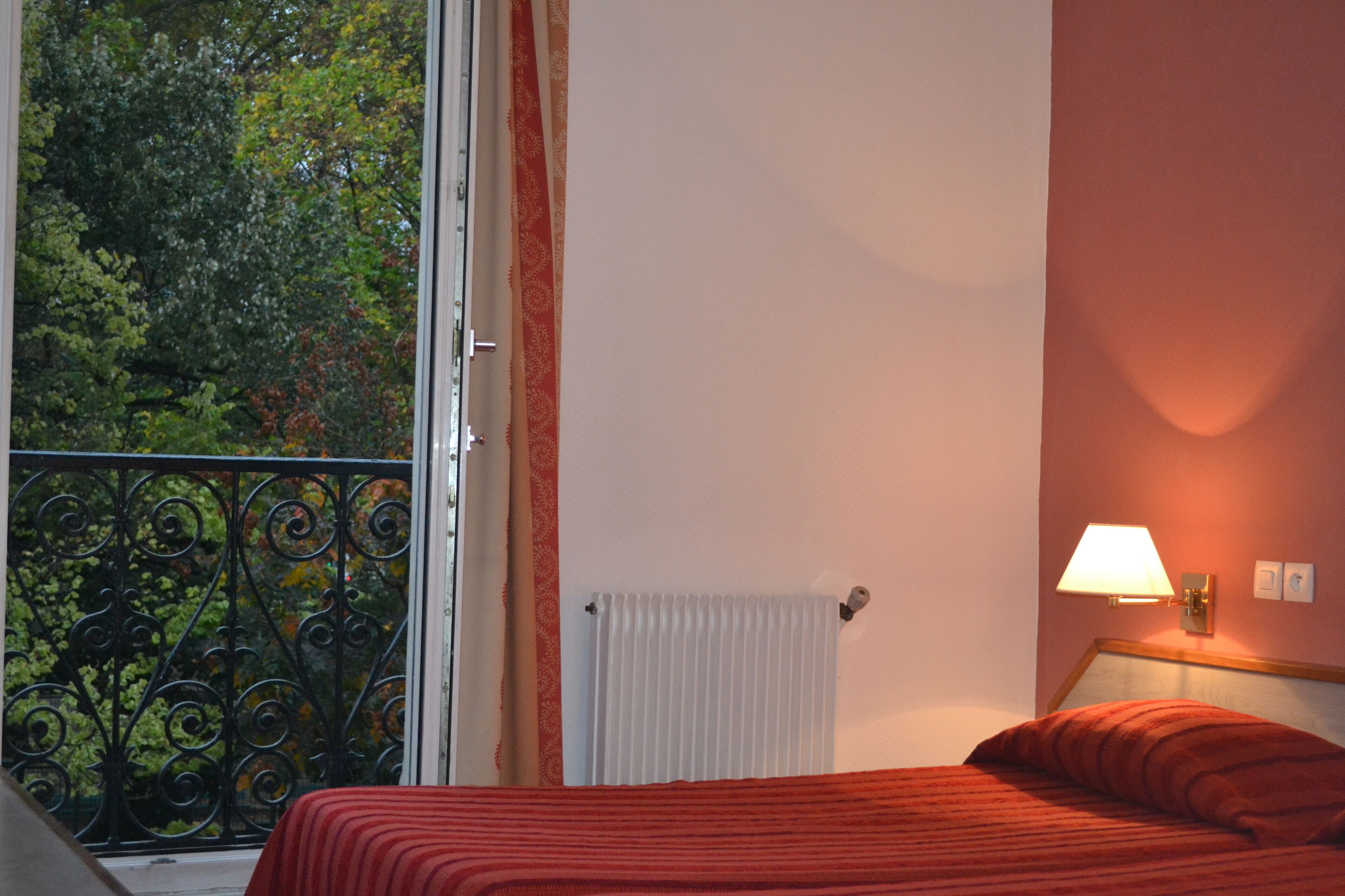 Un dormitorio con una cama, una ventana con vistas a árboles, un radiador y una lámpara fijada a la pared.