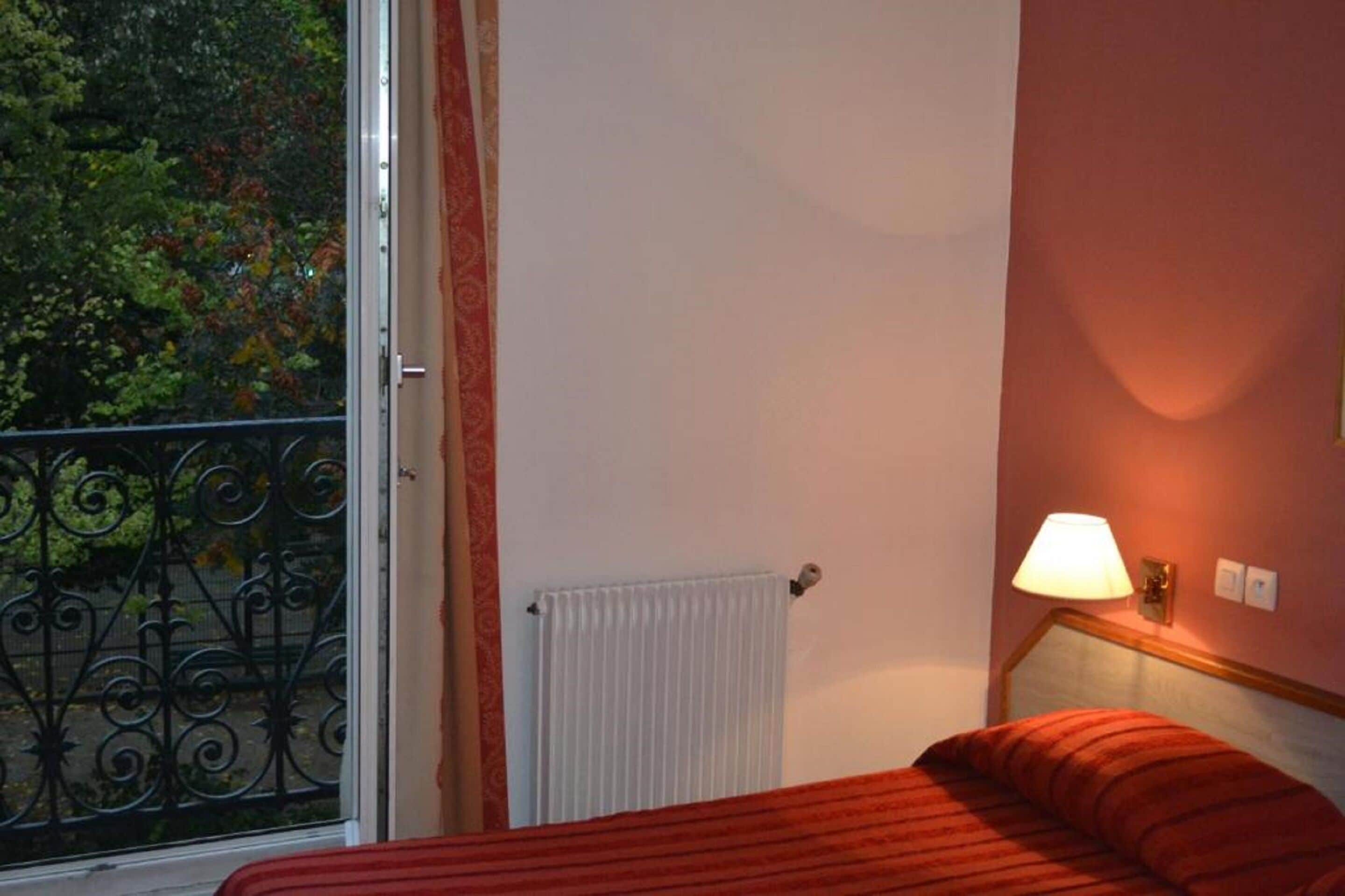 A hotel room with a bed, a radiator, a window with a view of trees, and a wall-mounted lamp.