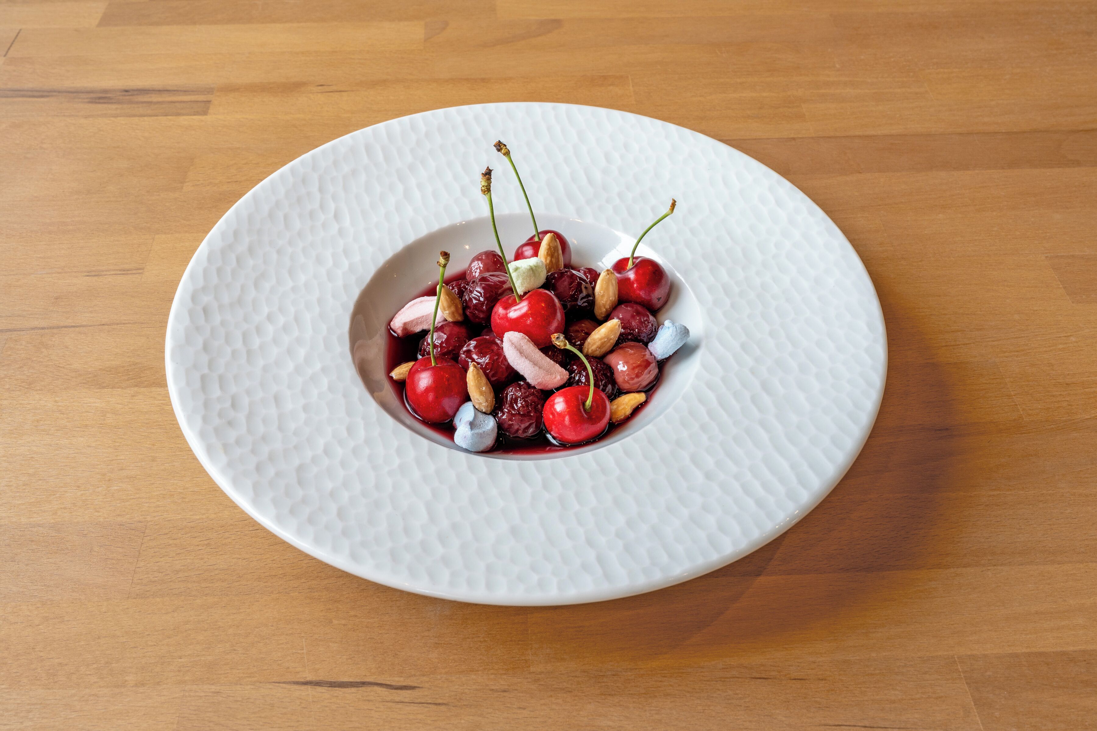 A white textured plate with a dessert of cherries, nuts, and possibly chocolate on a wooden table.