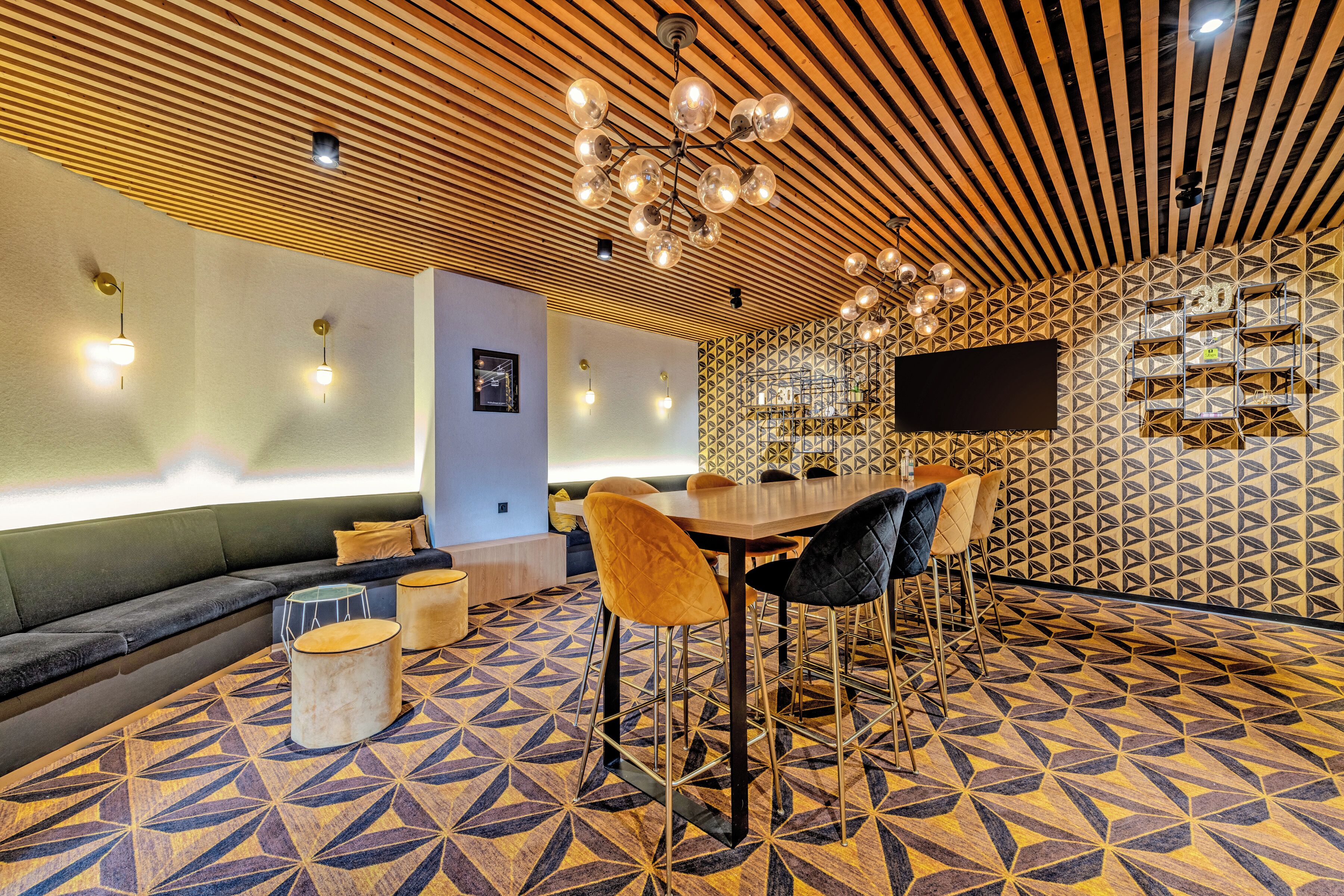 Un área de descanso moderna con suelo de patrón geométrico, techo de madera con luces colgantes, una mesa redonda con sillas y un televisor de pantalla plana montado en la pared.