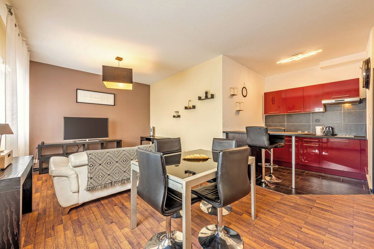 A modern living room with a sofa, dining table, and chairs, a TV, and a kitchen with red cabinets.