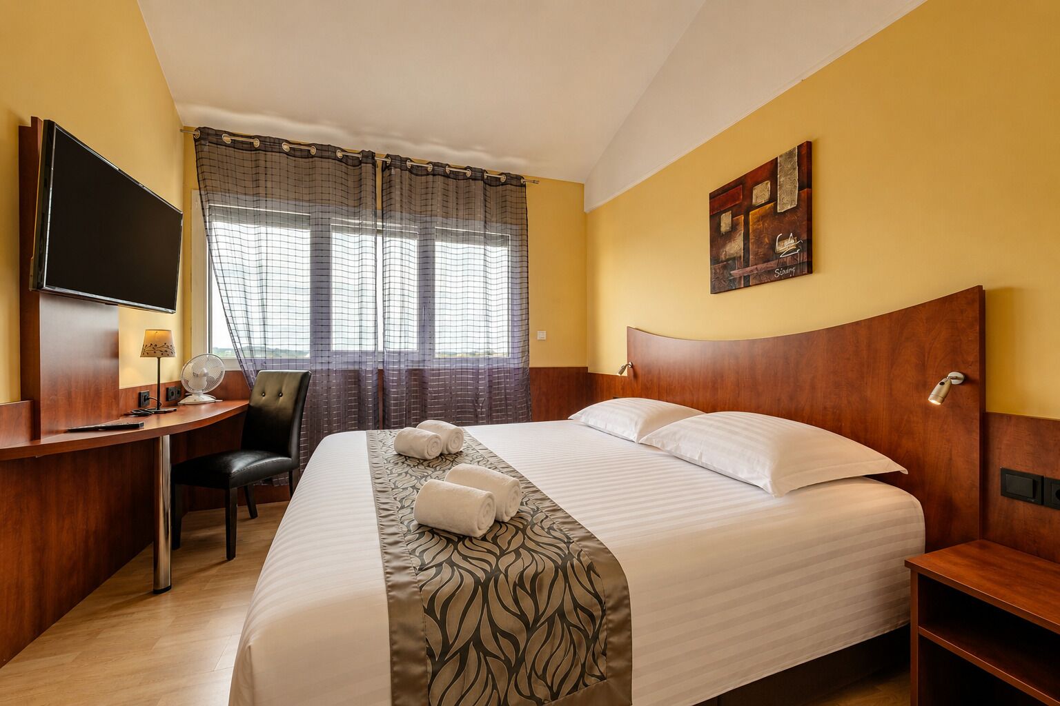 A hotel room with a bed, a desk with a TV, a chair, a window with curtains, and a painting on the wall.