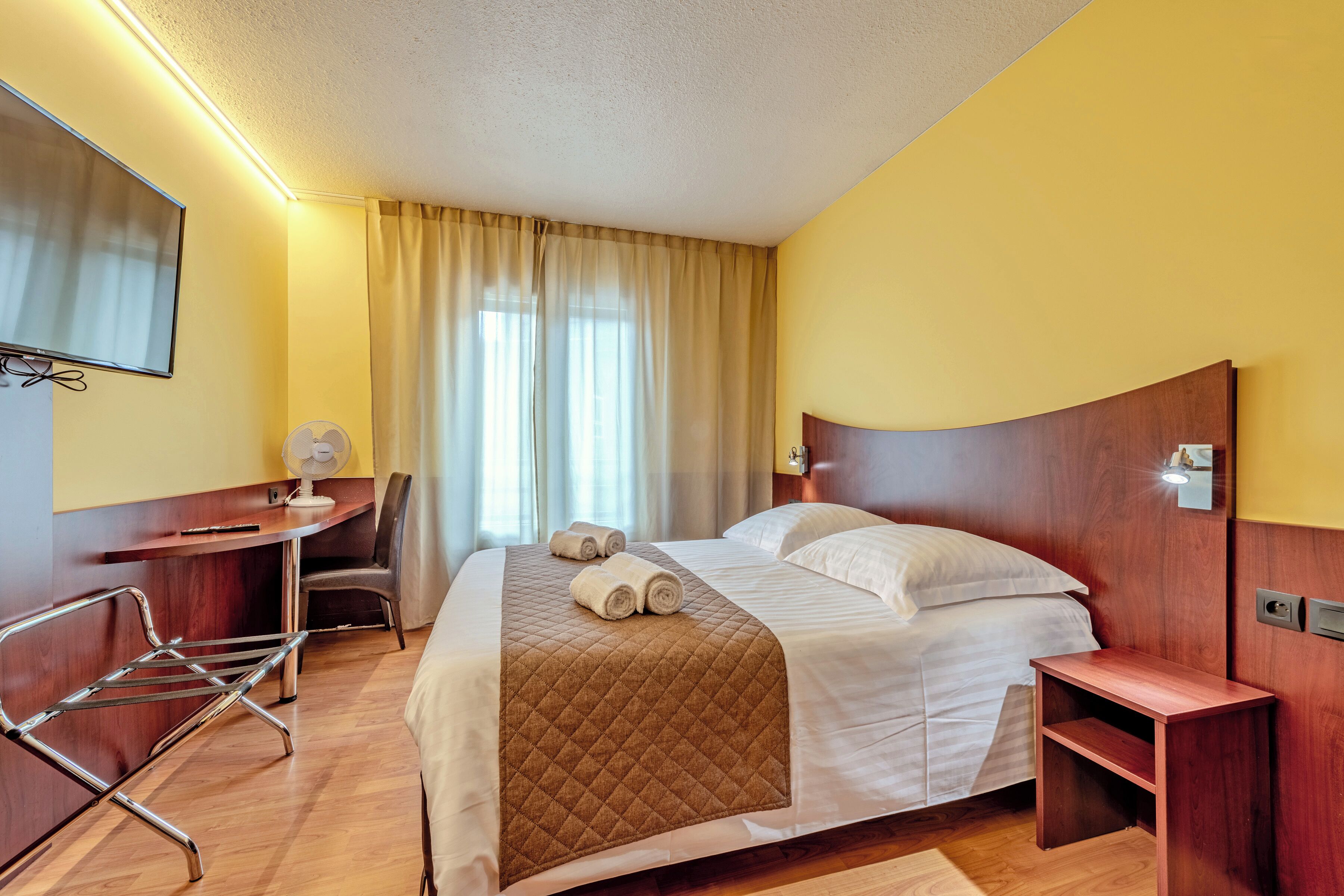 A hotel room with a bed, a desk, a chair, a TV, and a window with curtains.