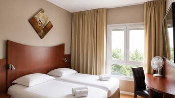 Ett hotellrum med två sängar, ett skrivbord, en stol, ett fönster med gardiner och ett konstverk som hänger på väggen.