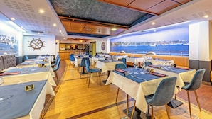 A restaurant with tables set for dining, a view of the sea, and a ship's wheel on the wall.