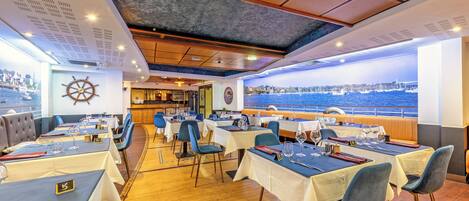 Un restaurant avec des tables dressées pour le dîner, une vue sur la mer et un gouvernail de bateau accroché au mur.