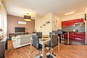 A modern living room with a sofa, dining table, and chairs, a flat-screen TV, and a kitchen with red cabinets.