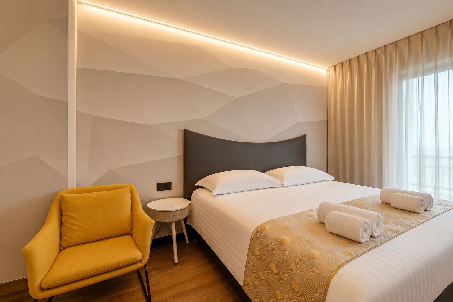 A hotel room with a bed, a round side table, a yellow armchair, and a window with curtains.