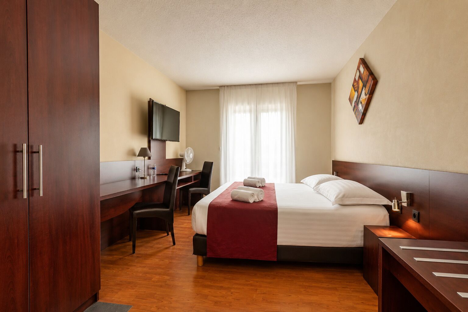 A hotel room with a bed, desk, chairs, a TV, and a window with curtains.