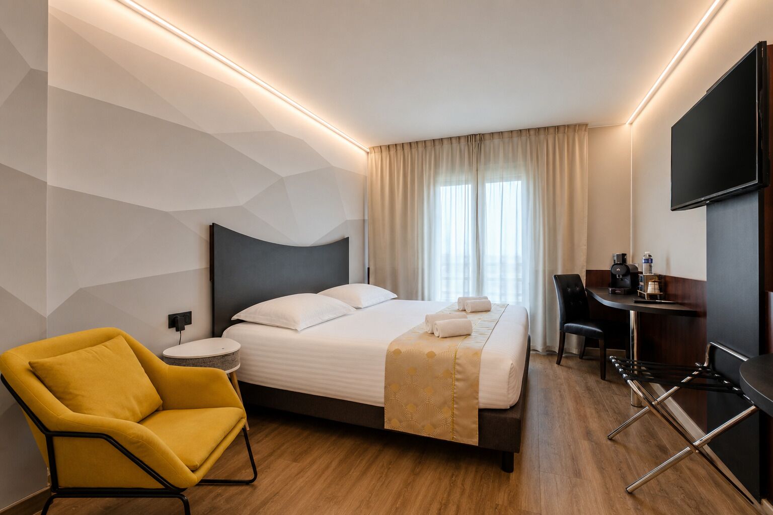 A modern hotel room with a bed, a desk, a chair, and a TV.