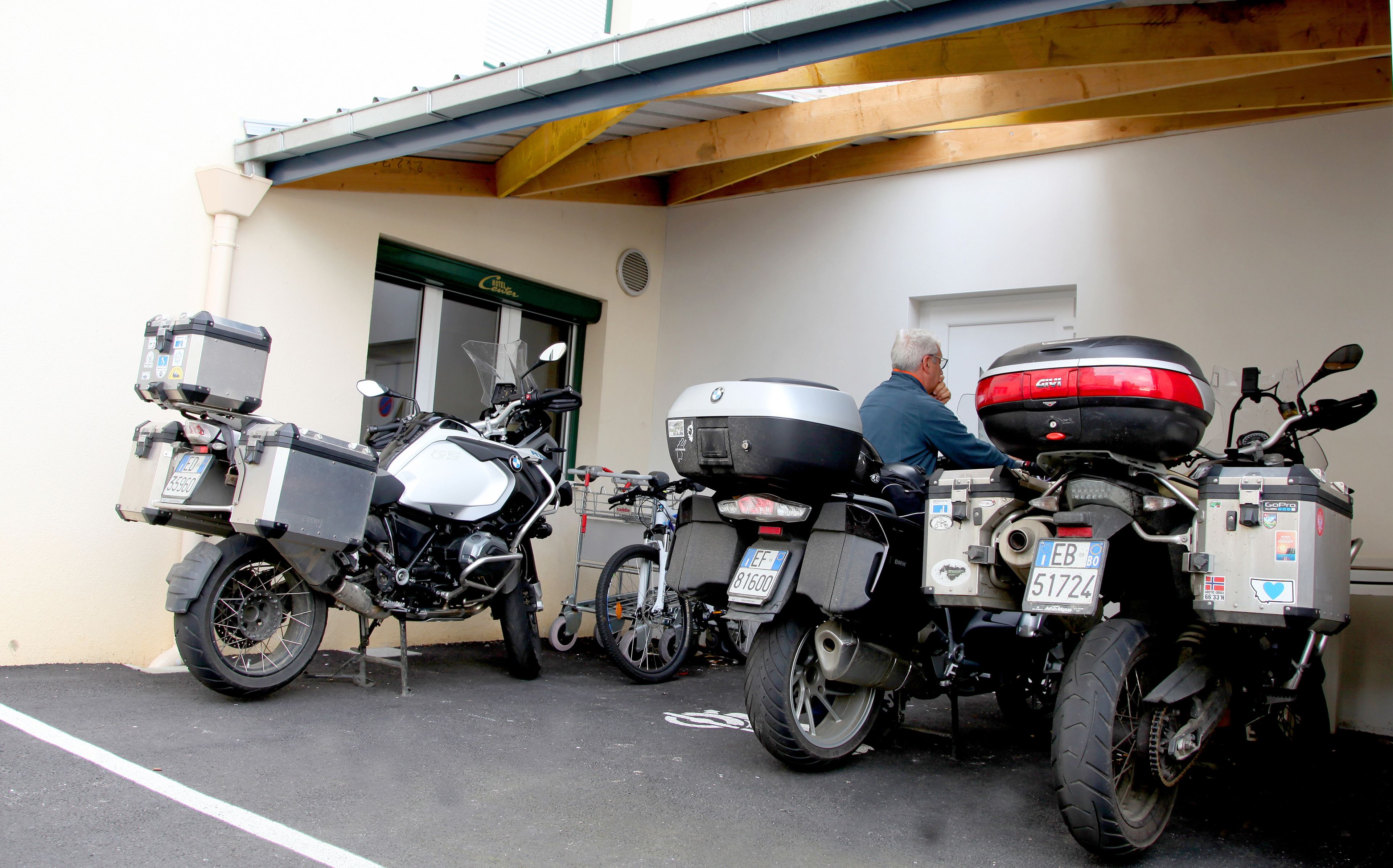 Tre motorcykler parkeret i et overdækket parkeringsområde med en bygning i baggrunden.