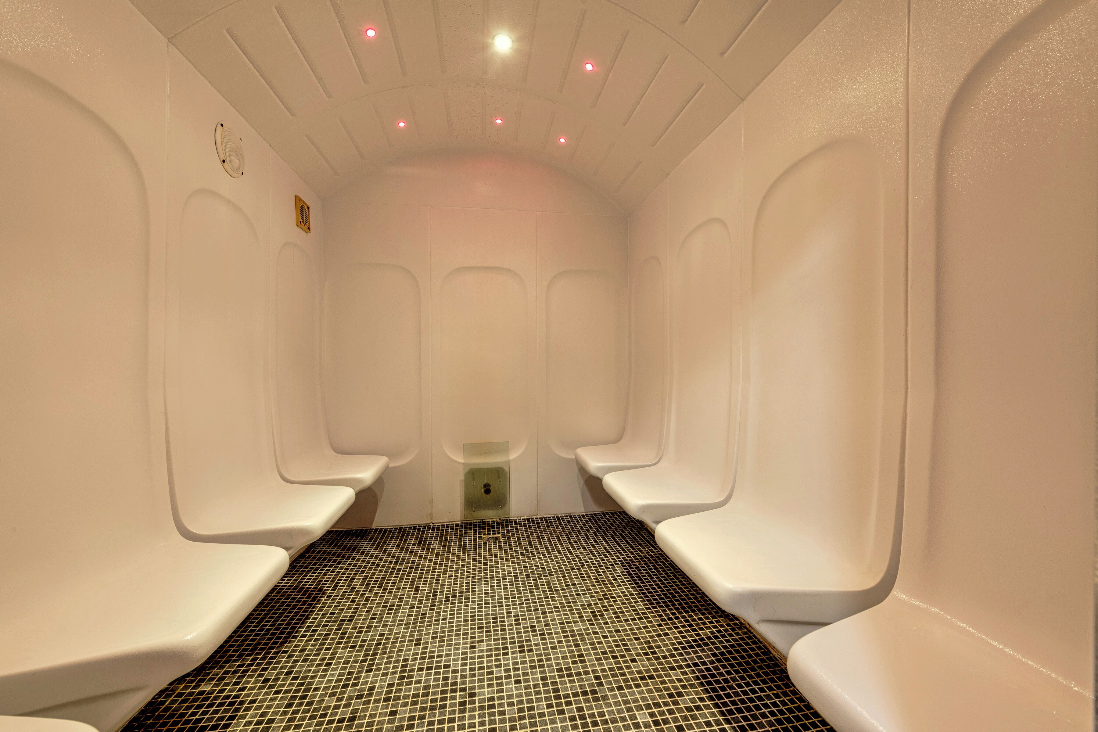 Une rangée de sièges blancs, de type banc, avec un sol carrelé, un plafond doté d’un éclairage encastré et une petite grille de ventilation.