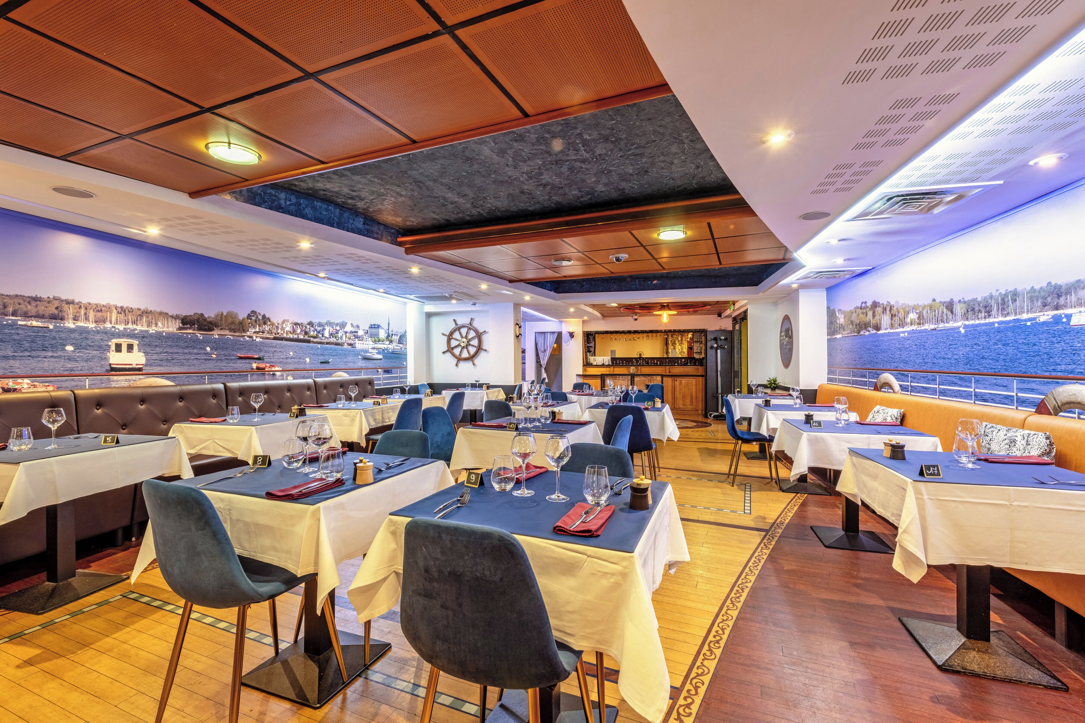 Een restaurant met tafels gedekt voor diners, uitzicht op zee en een scheepswiel aan de muur.