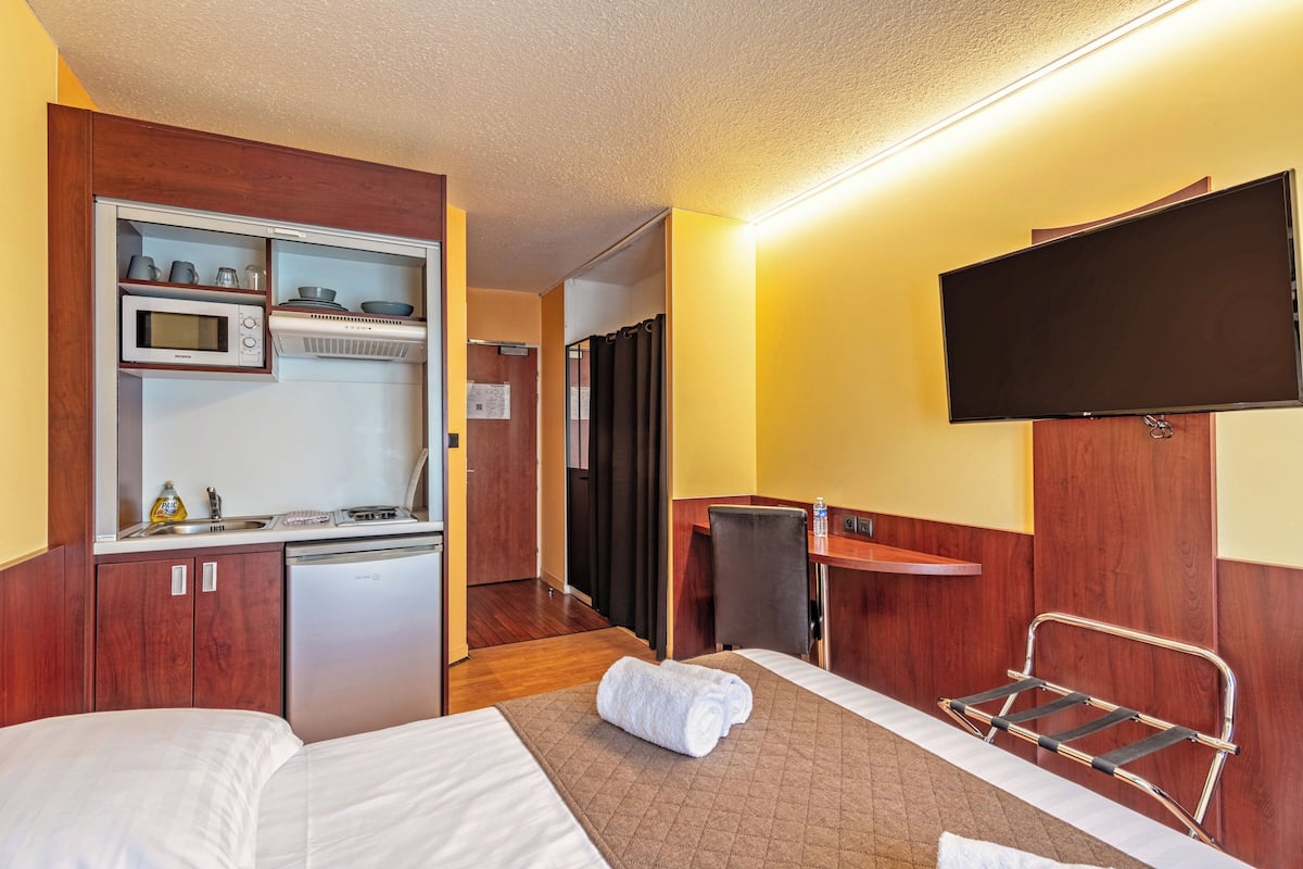 A hotel room with a kitchenette, a TV mounted on the wall, a bed with a rolled towel, and a chair.