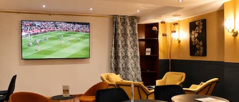 Ein Zimmer mit einem großen Fernseher, der ein Fußballspiel zeigt, gelben Stühlen, einem runden Tisch und einem Bücherregal.