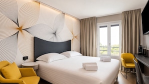 A hotel room with a large bed, two yellow chairs, a desk with a flat-screen TV, and a balcony with curtains.