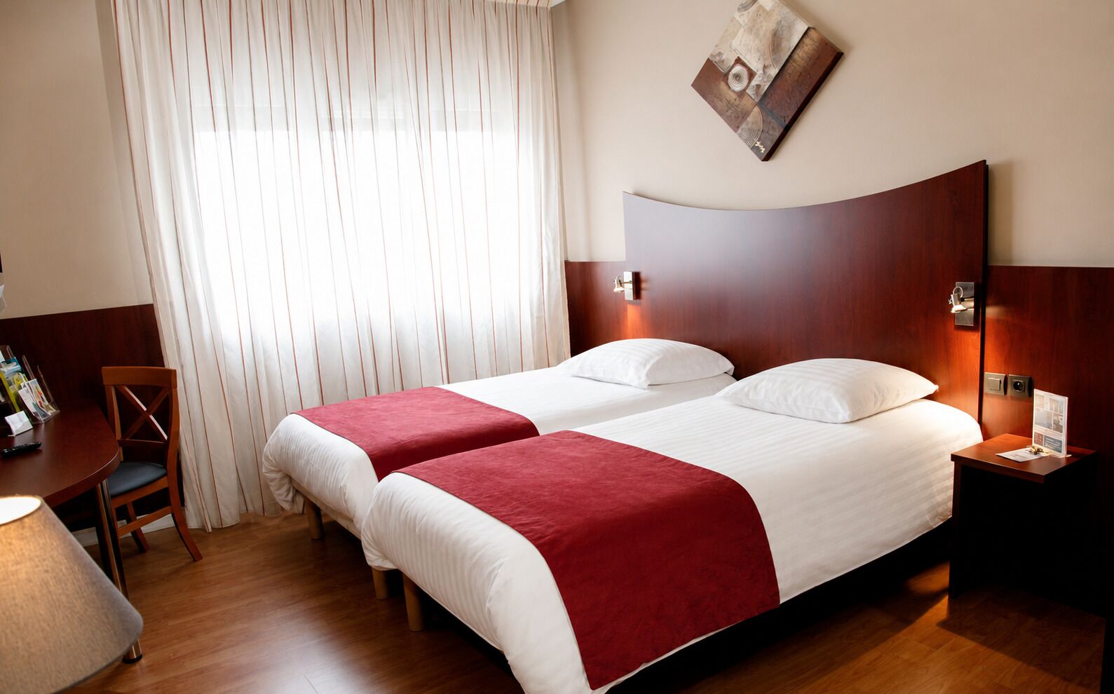 A hotel room with two beds, a wooden headboard, a desk with a chair, and a window with curtains.