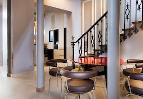 Un interior moderno con una escalera, una zona de comedor con mesas y sillas redondas, y una cocina a la vista.