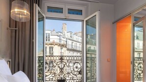 Una habitación de hotel con una cama grande, vistas de la ciudad a través de una puerta abierta y un balcón con barandilla de hierro forjado.