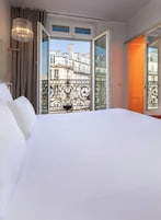 Una habitación de hotel con una cama grande, vistas de la ciudad a través de una puerta abierta y un balcón con barandilla de hierro forjado.