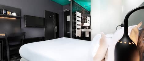 A modern hotel room with a bed, a desk, a TV, and a bathroom visible through a glass partition.