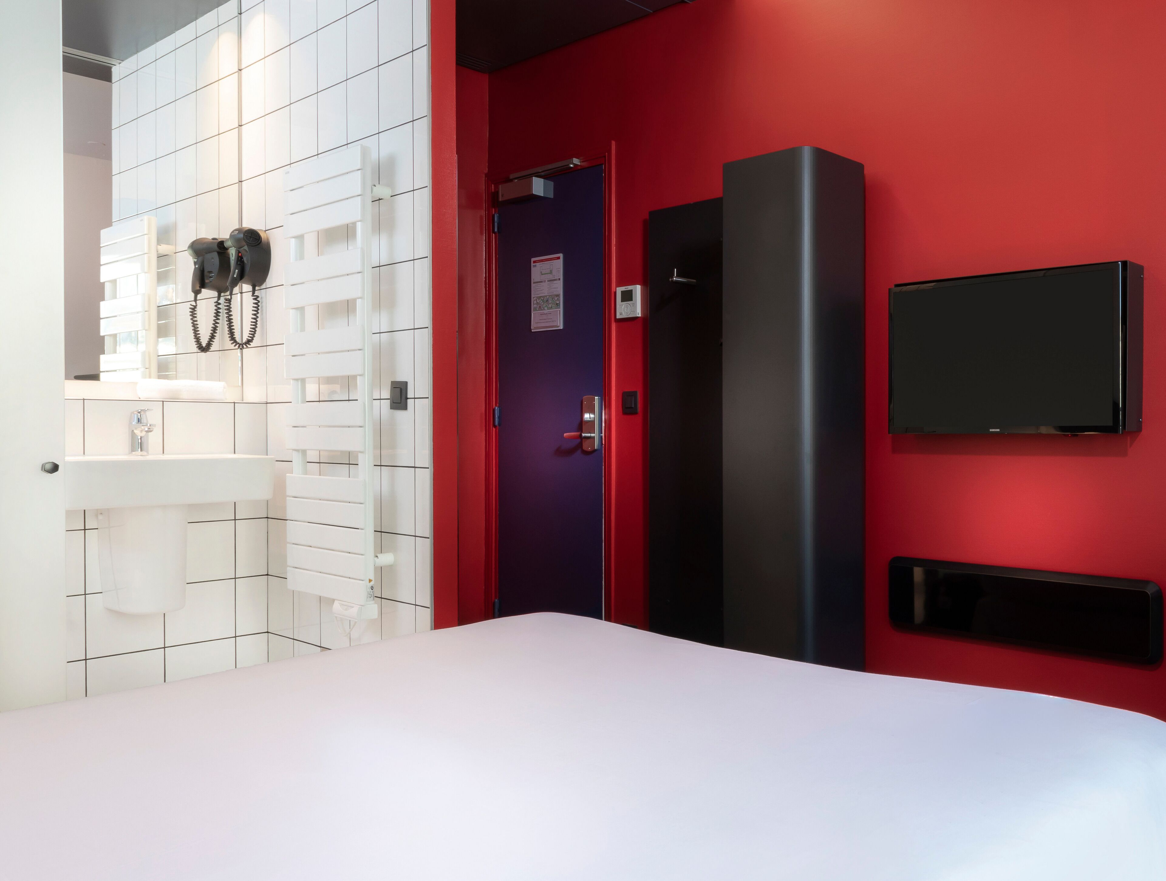A hotel room with a white bed, a red accent wall, a black TV, a white sink, and a door with a security lock.