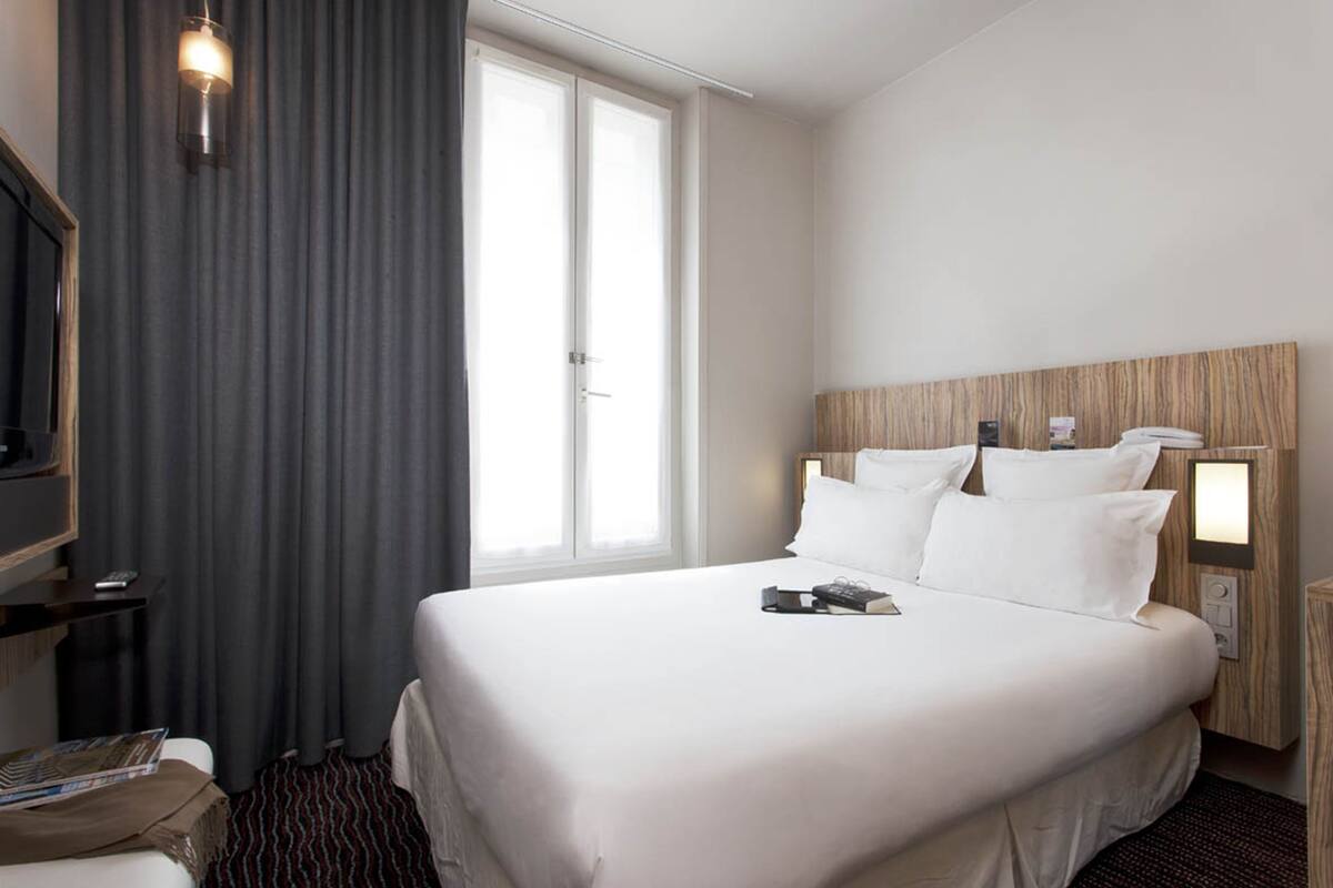 Habitación de hotel con una cama grande, una mesita de noche, una lámpara, una ventana con cortinas y un cabecero de madera.