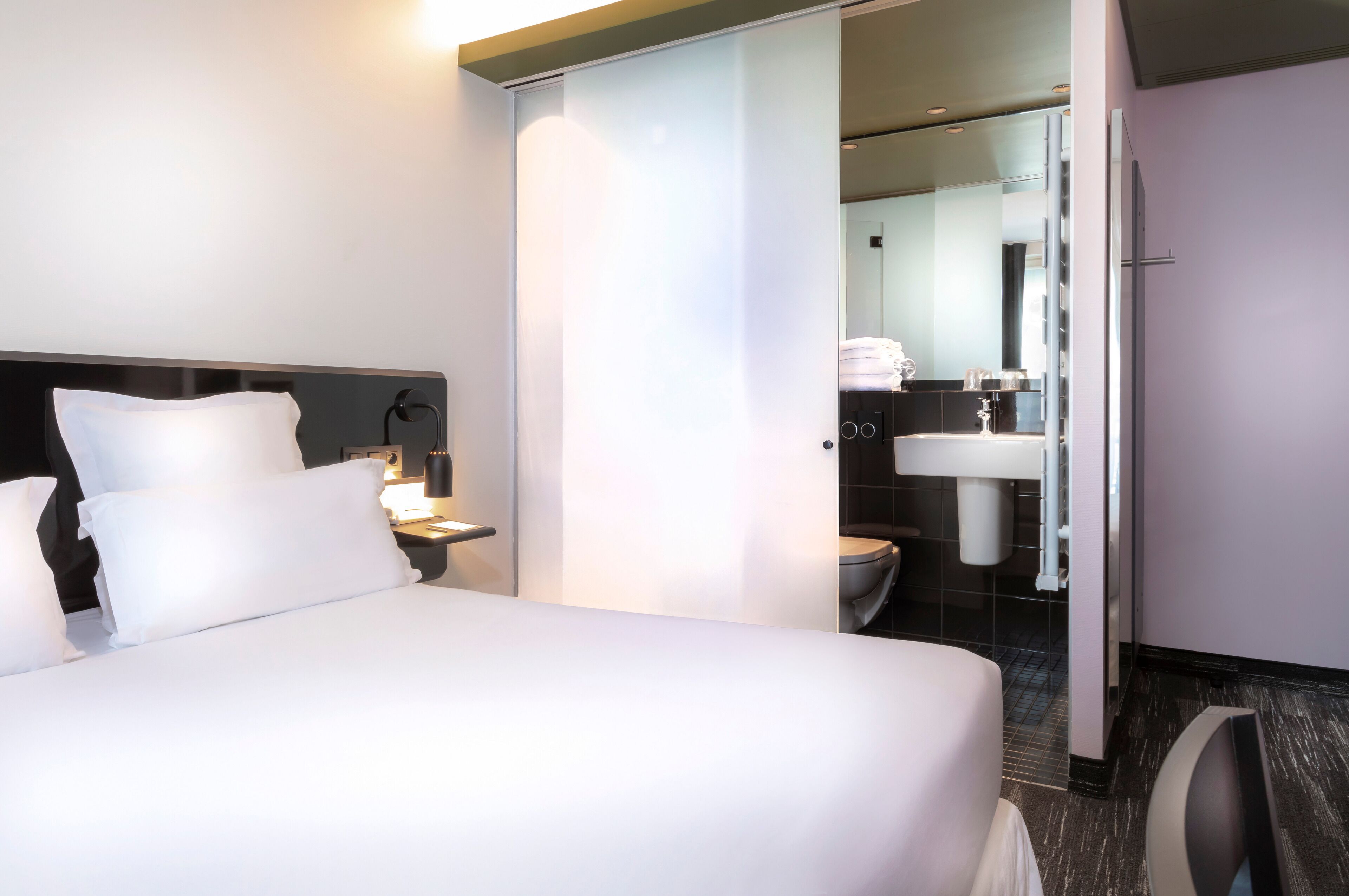 A hotel room with a bed, a nightstand, a mirror, and a bathroom visible through an open door.