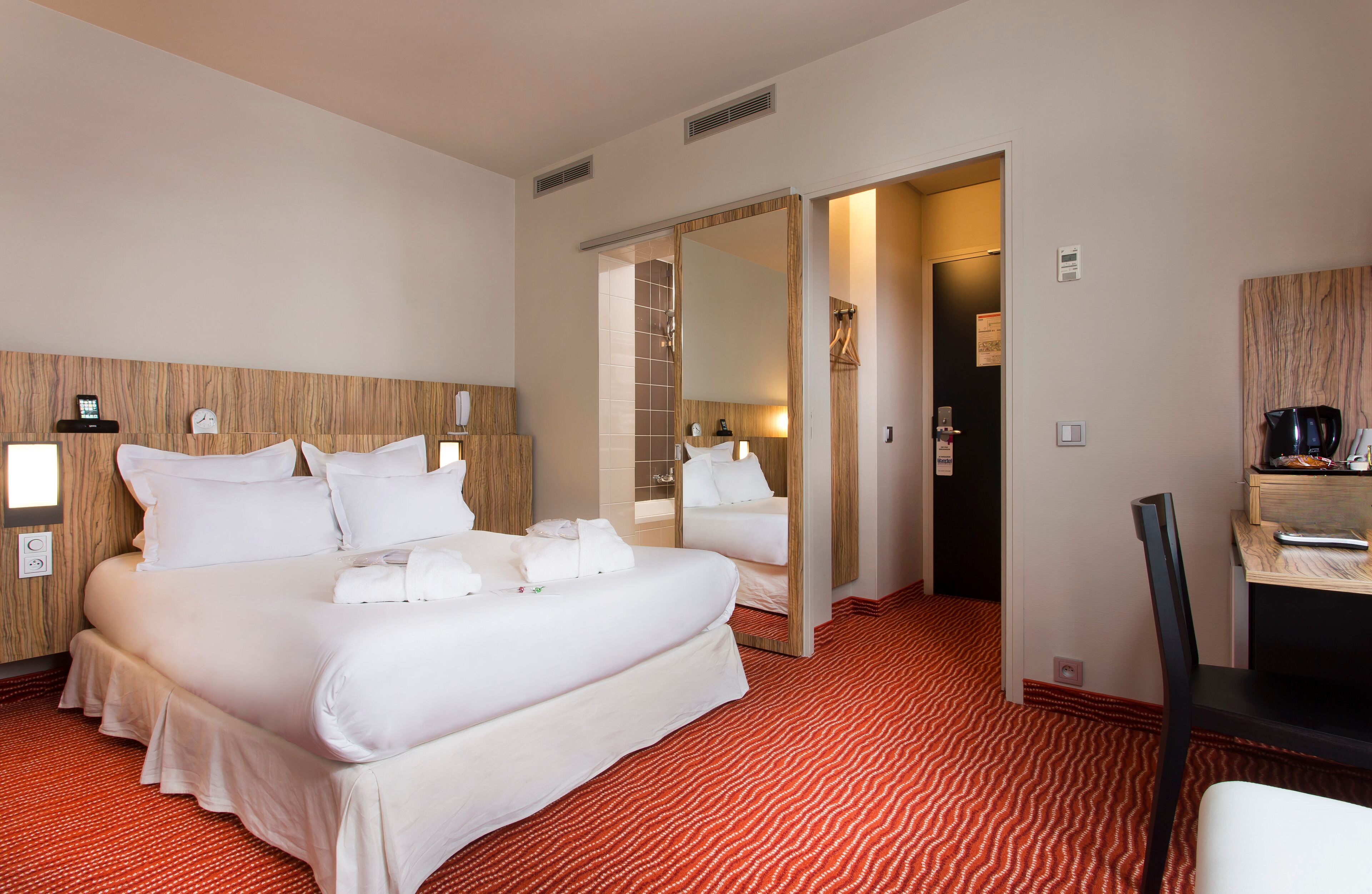 A hotel room with a large bed, a mirror, a desk, and a red patterned carpet.