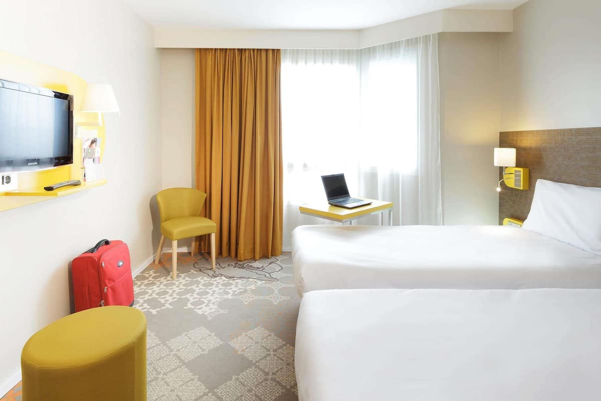 A hotel room with a bed, a desk with a laptop, a TV, a chair, a red suitcase, and a window with curtains.