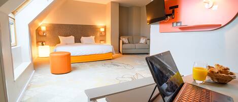 Una habitación de hotel moderna con una cama, un escritorio con una laptop, un televisor de pantalla plana y una bandeja de desayuno con croissants y zumo de naranja.